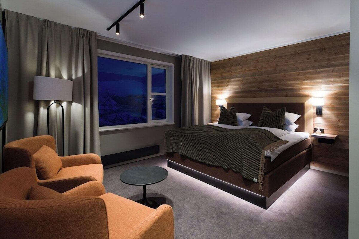 A modern hotel room with a large bed, a sitting area with a chair, a small table, and a window with curtains.