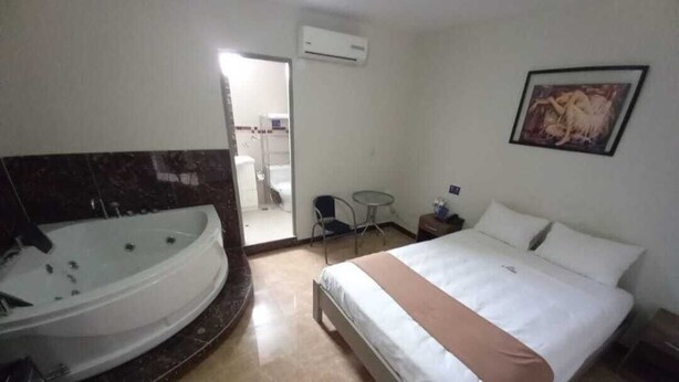 Una habitación de hotel con una bañera grande, una cama con ropa blanca, una mesita con una silla y un cuadro en la pared.