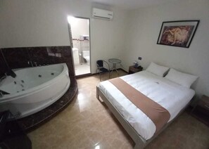 Una habitación de hotel con una bañera grande, una cama con ropa blanca, una mesita con una silla y un cuadro en la pared.