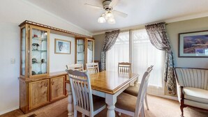 Comedor con mesa de madera, sillas blancas, aparador con puertas de vidrio, ventilador de techo y ventana con cortinas.