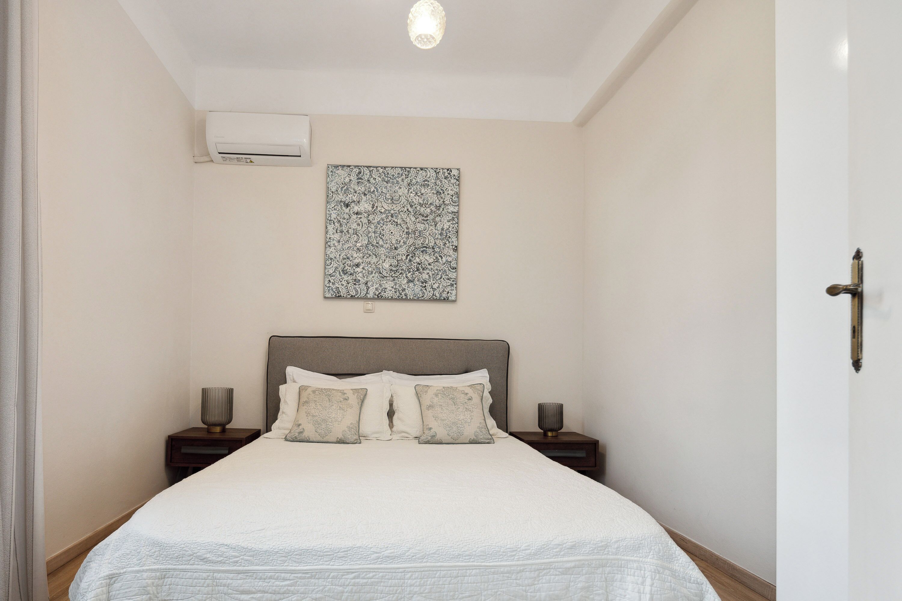 A neatly made bed with a white duvet and grey headboard, two bedside tables with lamps, a wall-mounted air conditioner, and a patterned artwork above the bed.