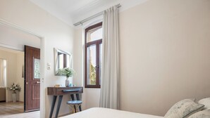 Une chambre avec un grand lit, un bureau avec une chaise, une fenêtre avec des rideaux et une porte donnant sur une autre pièce.