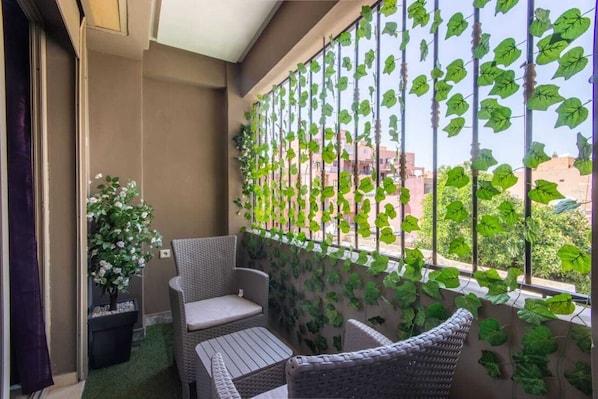 A balcony with wicker furniture, a potted plant, and a view of buildings through a metal fence with green foliage.