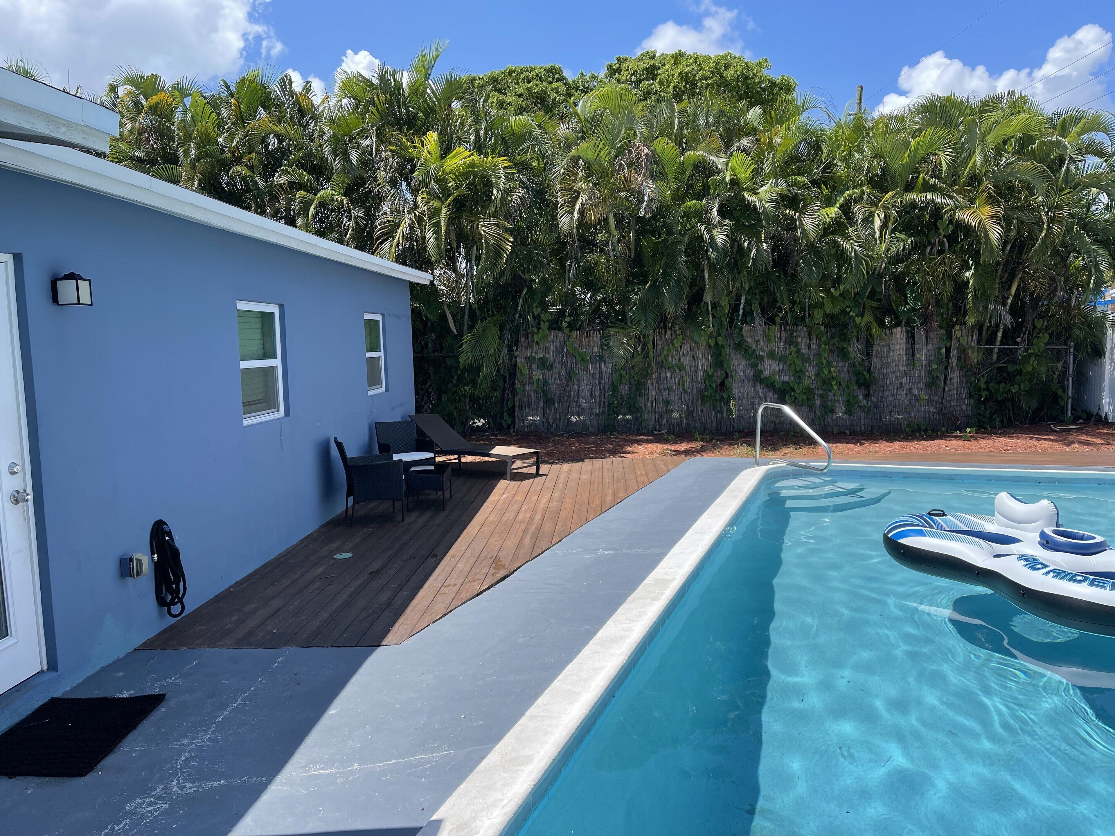 Un área de piscina con una casa azul, sillas de descanso y una terraza de madera.
