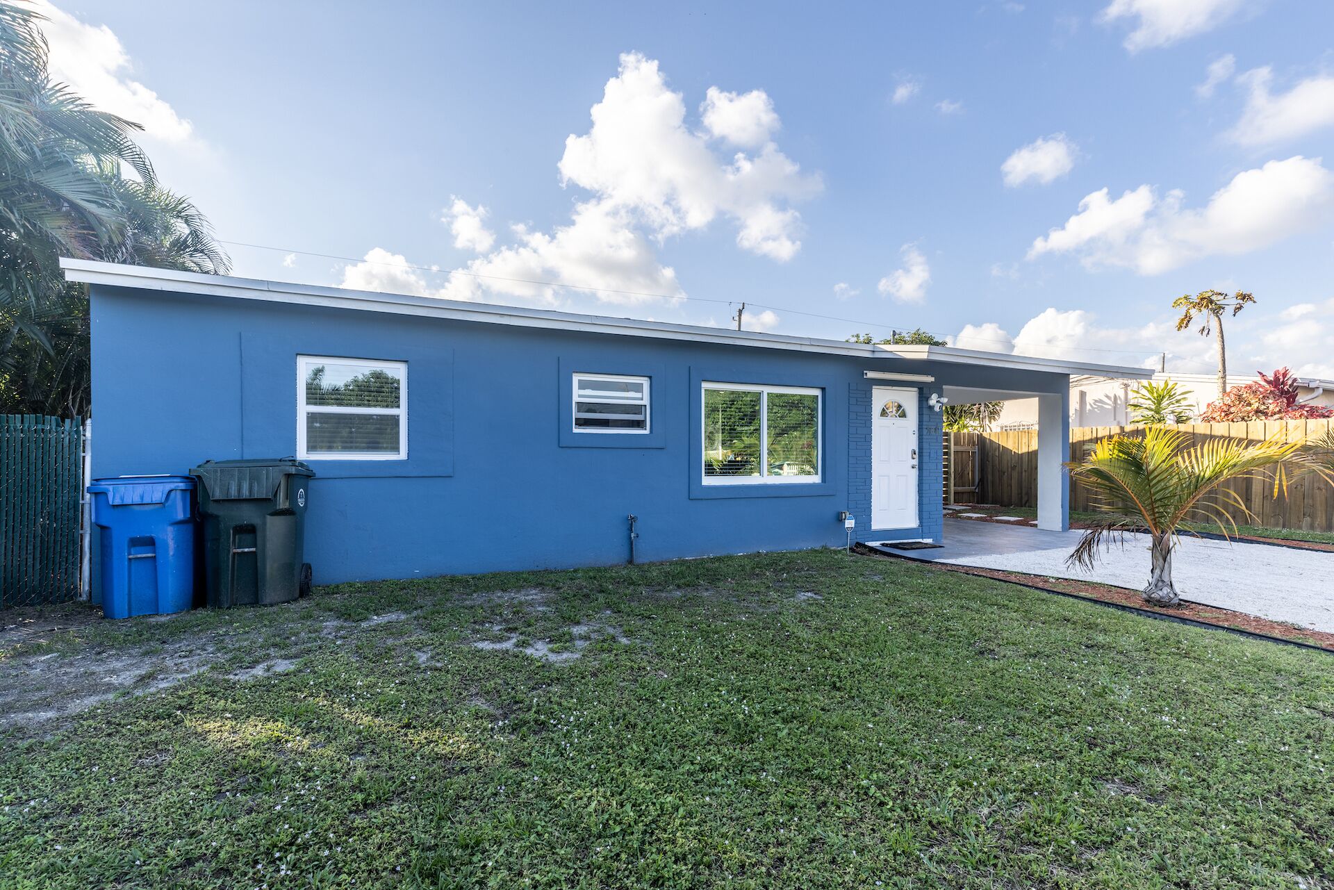 A single-story blue house with a front door, two windows, and a small front yard with grass and a palm tree.