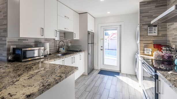 A modern kitchen with white cabinetry, a granite countertop, stainless steel appliances, and a view of the outside. - Beach Oasis with Private Pool (Fort Lauderdale)