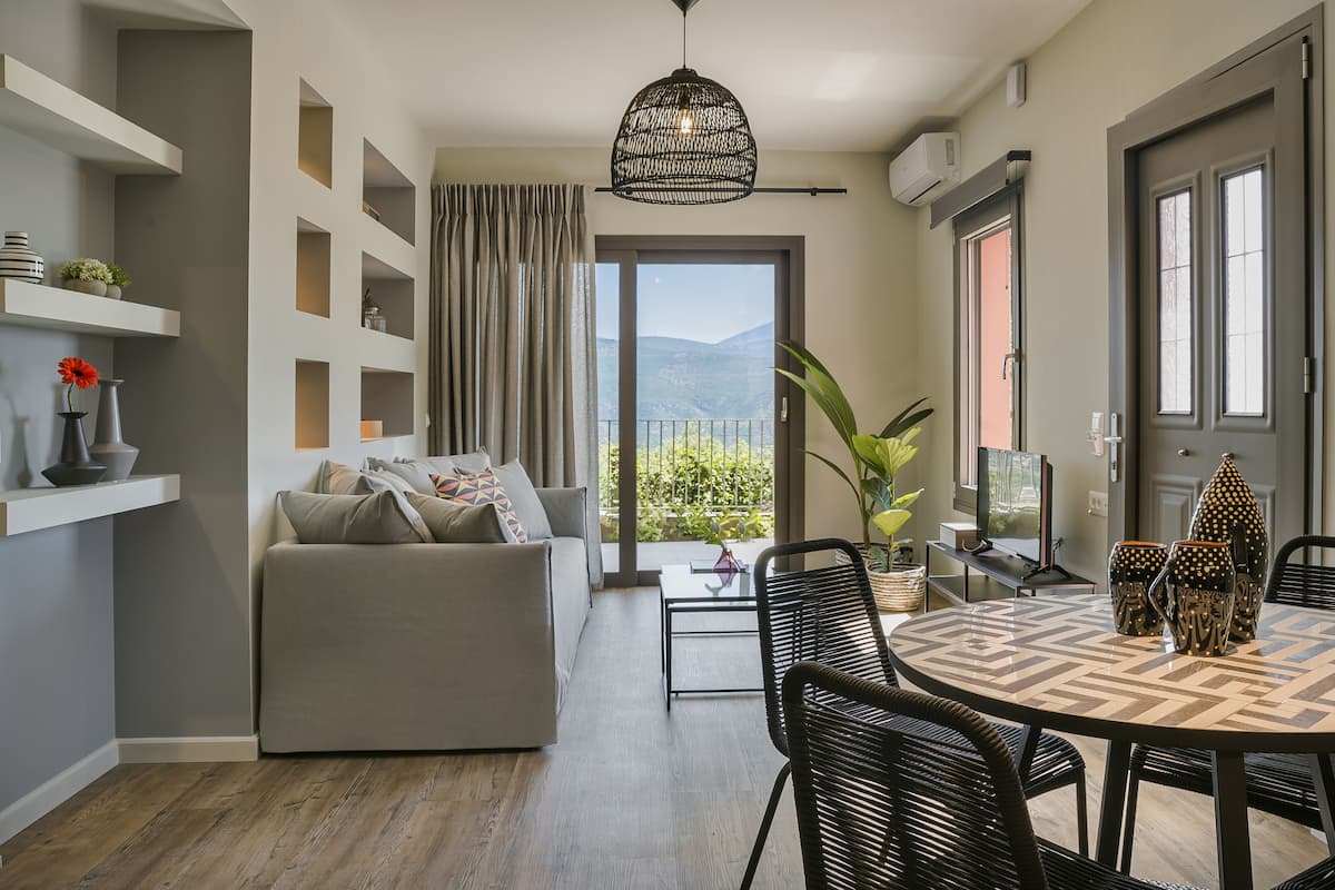 A modern living room with a sofa, dining table, and chairs, a large window with a view of mountains, and a hanging light fixture.