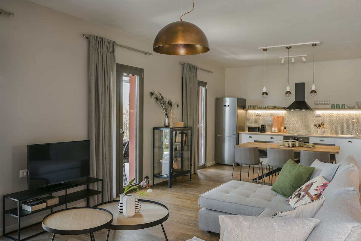A modern living room with a flat-screen TV, a sofa, a coffee table, and a dining area with a kitchen in the background.