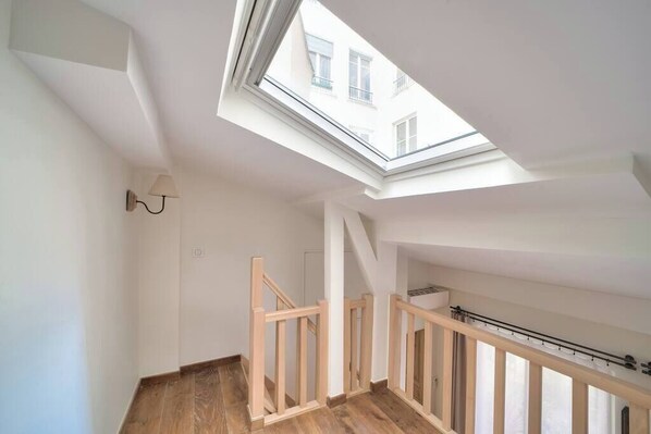 Une pièce aux hauts plafonds, dotée d’une verrière, d’un escalier en bois et offrant une vue sur les bâtiments environnants.