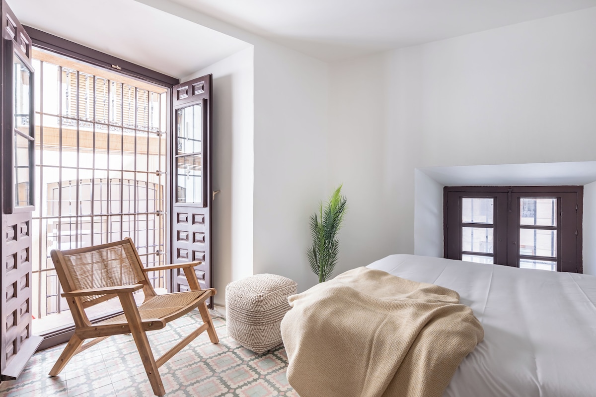 A bedroom with a bed, a wooden chair, a small round ottoman, a potted plant, and a window with a view of buildings.