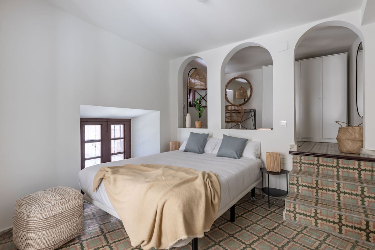 Quarto com uma cama, um banquinho, um banco, um espelho e piso estampado.