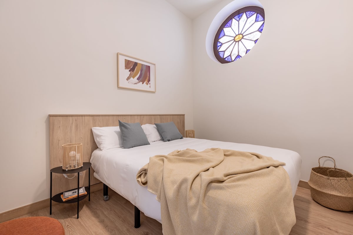 Um quarto com cama, mesa de cabeceira com abajur, janela com detalhe em vitral e um quadro na parede.