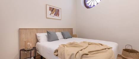 A bedroom with a bed, a bedside table with a lamp, a window with a stained glass design, and a framed picture on the wall.