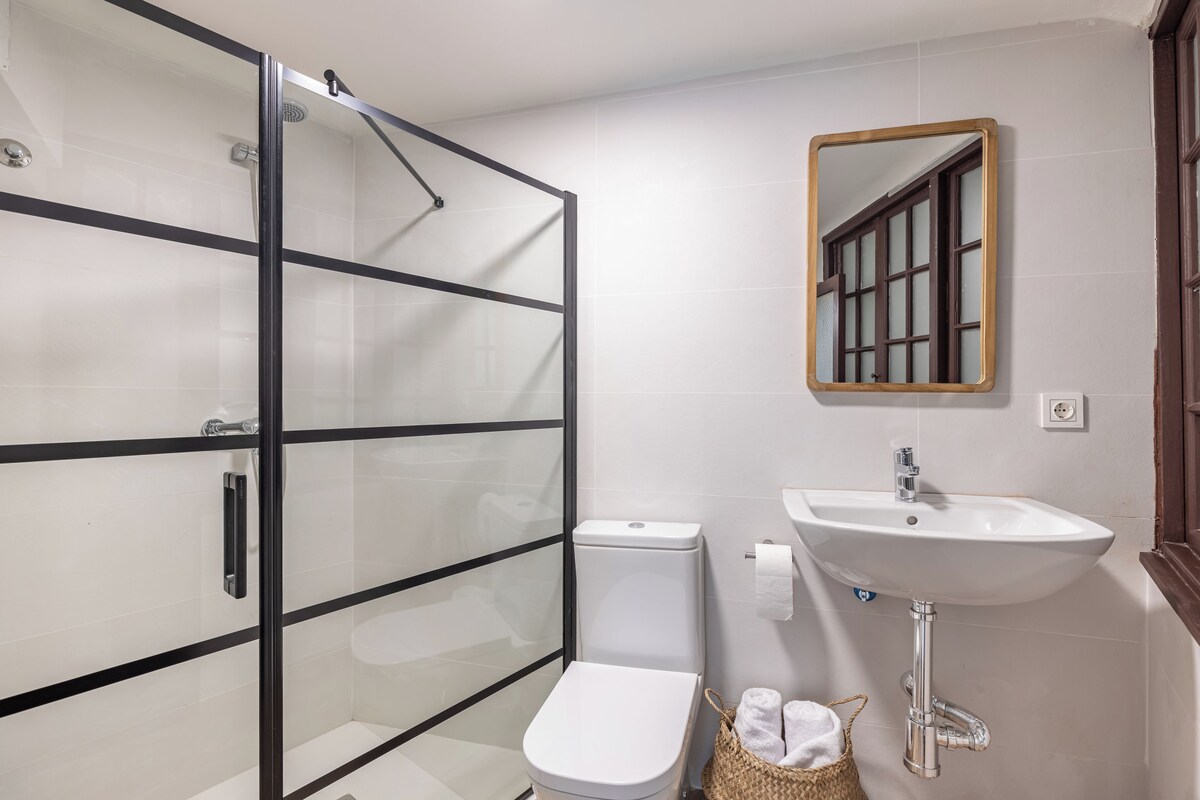 Un baño con cabina de ducha de vidrio, inodoro blanco, lavabo con espejo y una cesta con toallas.