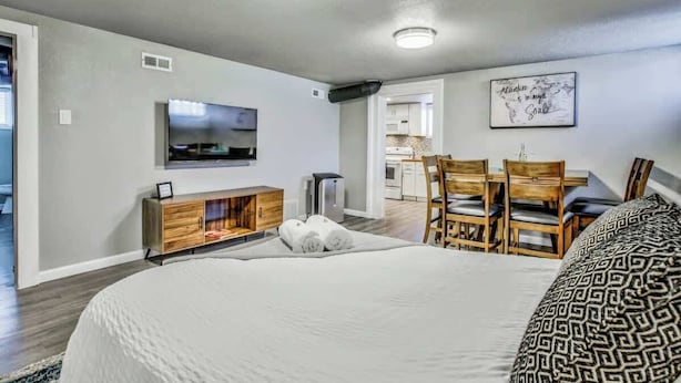 Un dormitorio moderno con una cama grande, un televisor montado en la pared, un mueble de madera y un rincón de comedor con sillas.