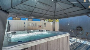 Un jacuzzi bajo un techo de metal, con una terraza de madera y un muro azul al fondo.