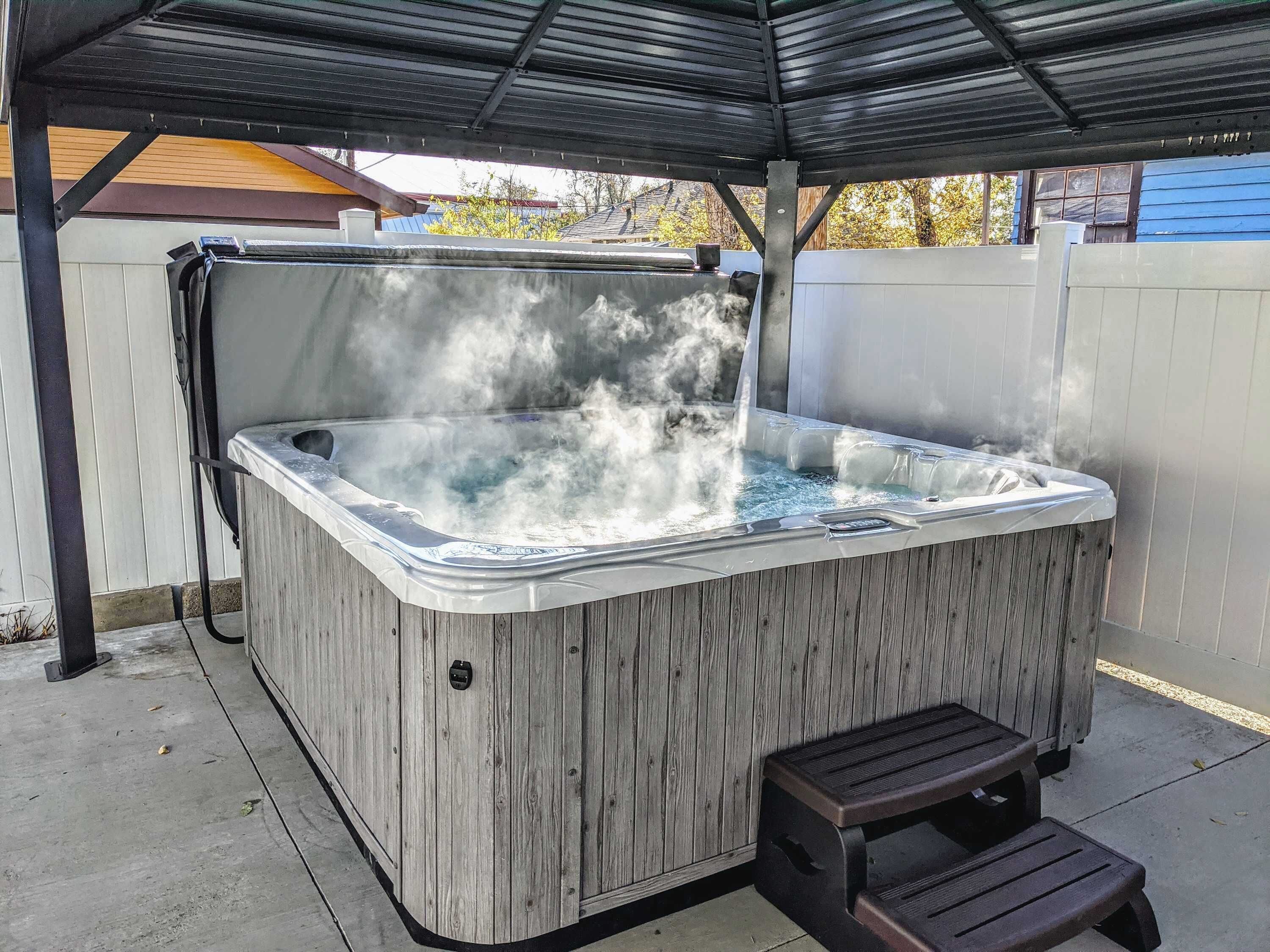 A hot tub with a wooden exterior and a metal roof, positioned on a concrete patio with a wooden bench nearby.