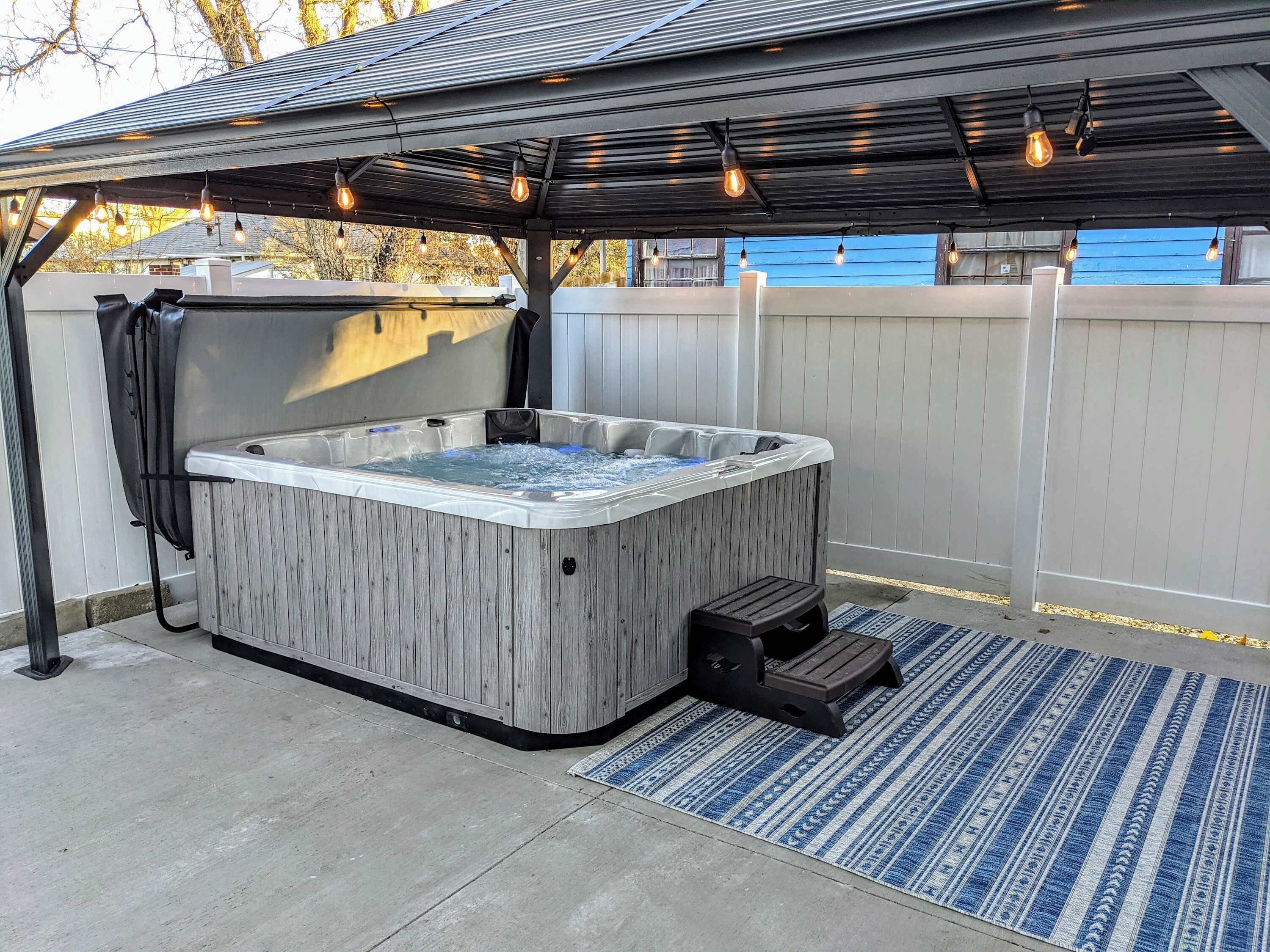 Un bain à remous sous une pergola illuminée de guirlandes lumineuses, entouré d’une clôture en bois et agrémenté d’un tapis bleu à motifs.