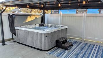 Un bain à remous sous une pergola illuminée de guirlandes lumineuses, entouré d’une clôture en bois et agrémenté d’un tapis bleu à motifs.