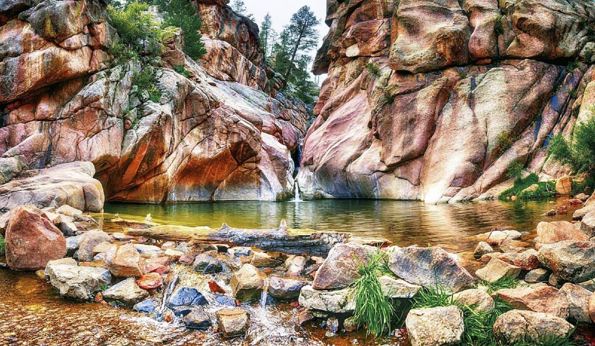 Eine natürliche Landschaft mit einem Wasserfall, felsigem Gelände und einem ruhigen Wasserbecken, umgeben von großen Felsen und viel Grün.