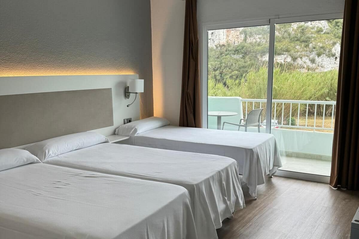 A hotel room with two beds, a balcony, and a view of greenery.