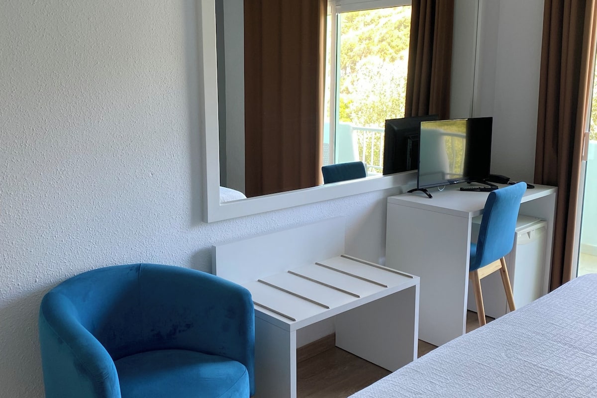 A hotel room with a bed, a desk with a computer, a blue chair, and a window with curtains.