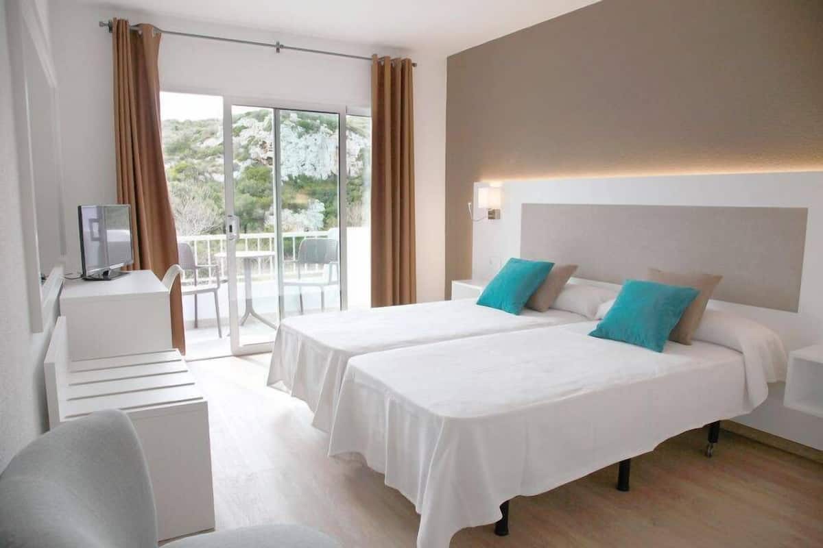 A hotel room with two beds, a desk, and a balcony with a view of greenery.