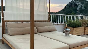 Un lit à baldaquin en bois, avec une literie beige et un ciel de lit, installé sur un balcon offrant une vue sur un paysage montagneux.
