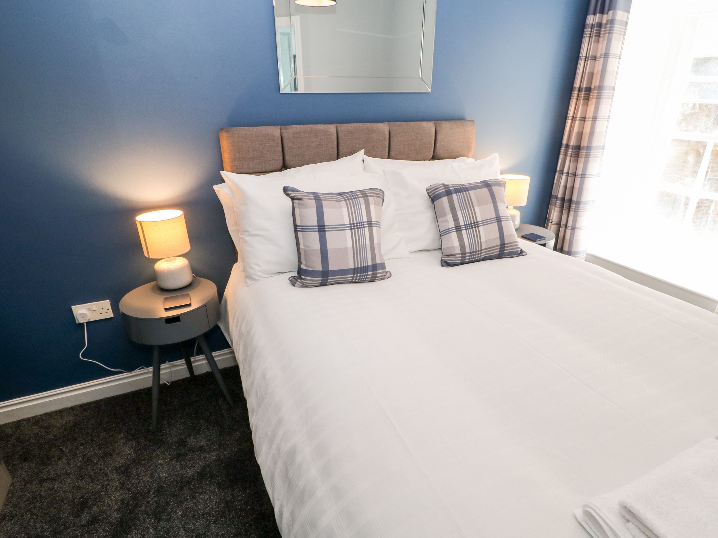 A neatly made bed with white linens and two plaid pillows, a bedside lamp, and a small table with a lamp.