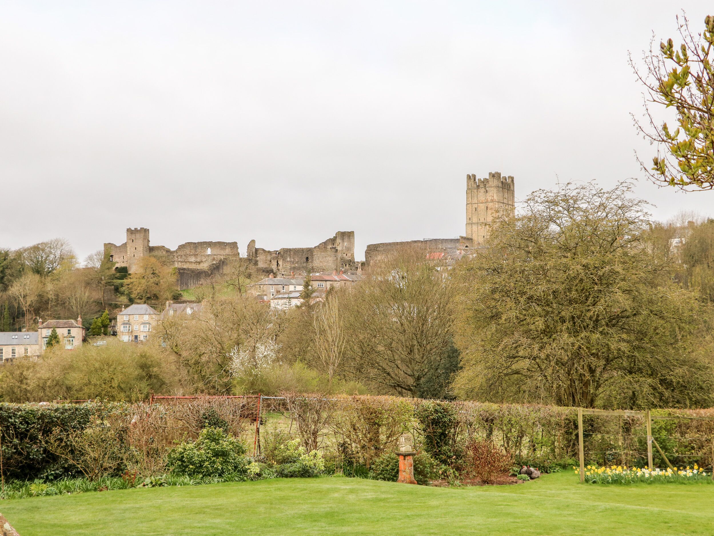 A castle with ruins and a tower, surrounded by trees and a grassy area with a hedge and a statue.