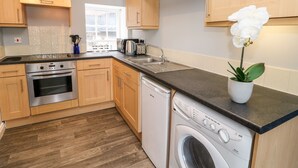 A modern kitchen with wooden cabinets, a stainless steel stove, a dishwasher, and a washing machine. - 3 Kings Mews (Richmond)