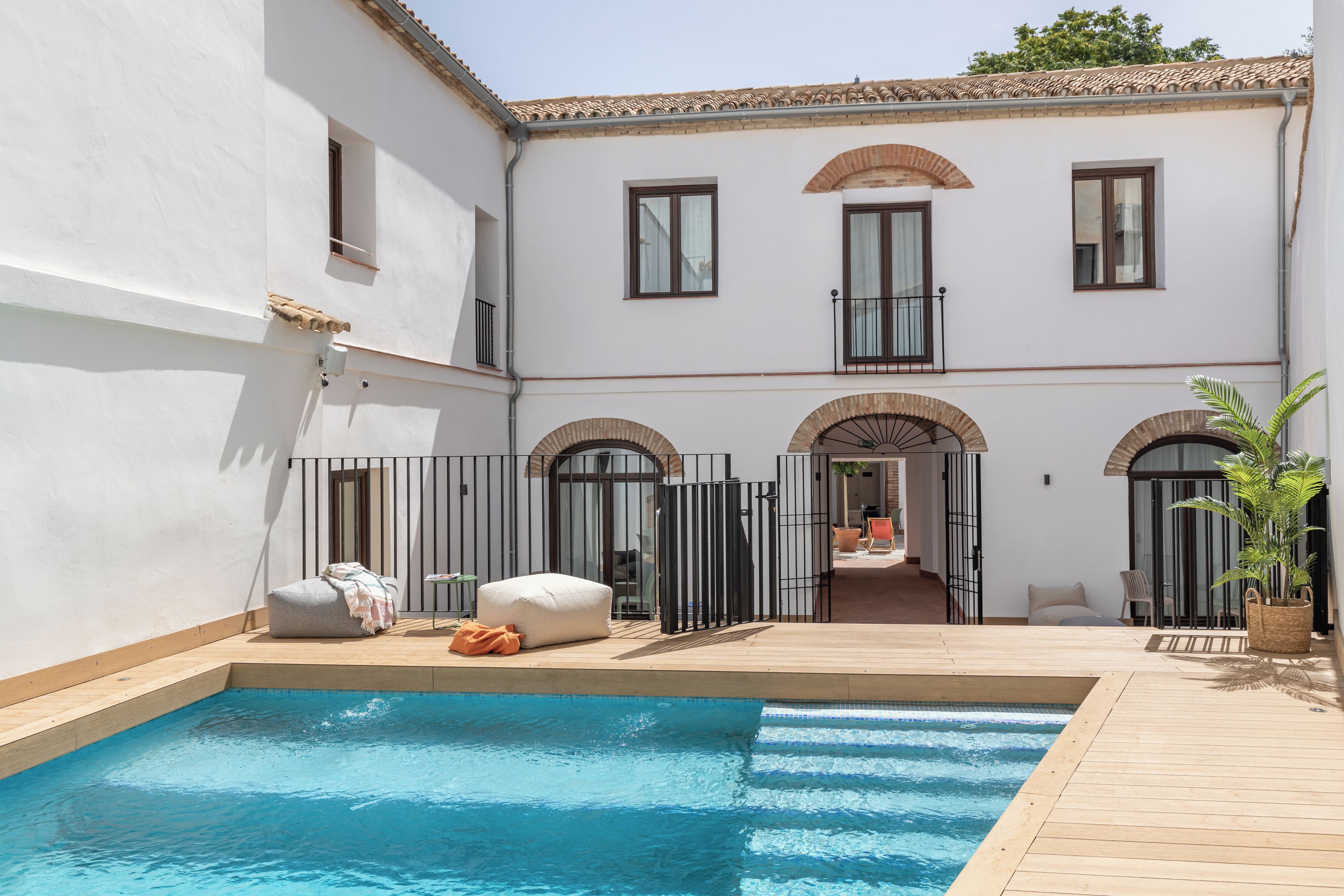 A poolside area with a wooden deck, a white building with arched windows, and a covered seating area.