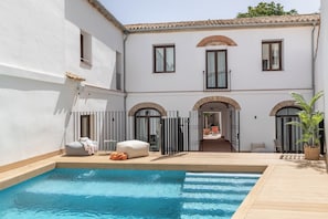 Un área junto a la piscina con cubierta de madera, un edificio blanco con ventanales arqueados y una zona de estar techada.