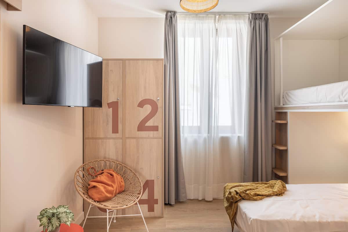 Een moderne hotelkamer met een flatscreen televisie, een houten garderobe met cijfers, een rieten stoel, een bed met gouden plaid en een groot raam met gordijnen.