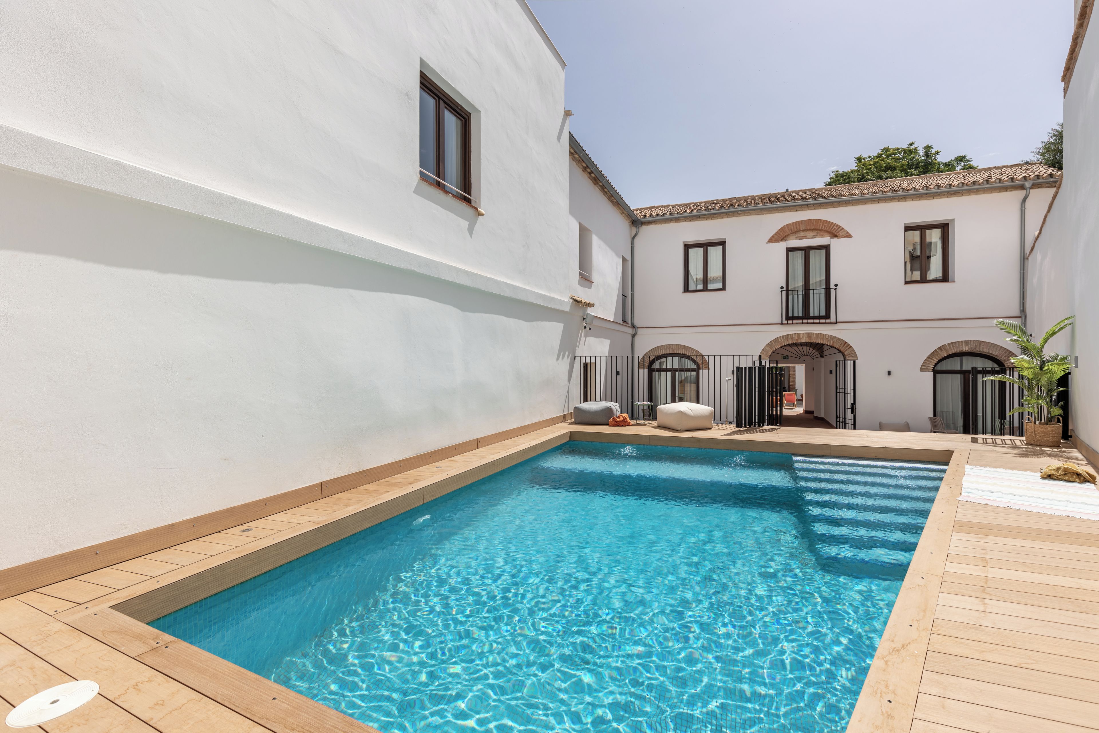 A swimming pool with a wooden deck, surrounded by a white building with arched windows and a balcony.