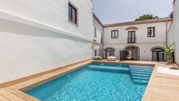 A swimming pool with a wooden deck, surrounded by a white building with arched windows and a balcony.