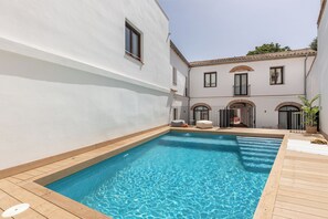 Una piscina con plataforma de madera, rodeada por un edificio blanco con ventanas arqueadas y un balcón.