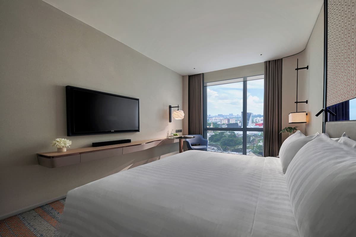 A modern hotel room with a large bed, a flat-screen TV mounted on the wall, a small table with a vase of flowers, and a view of the city through large windows.