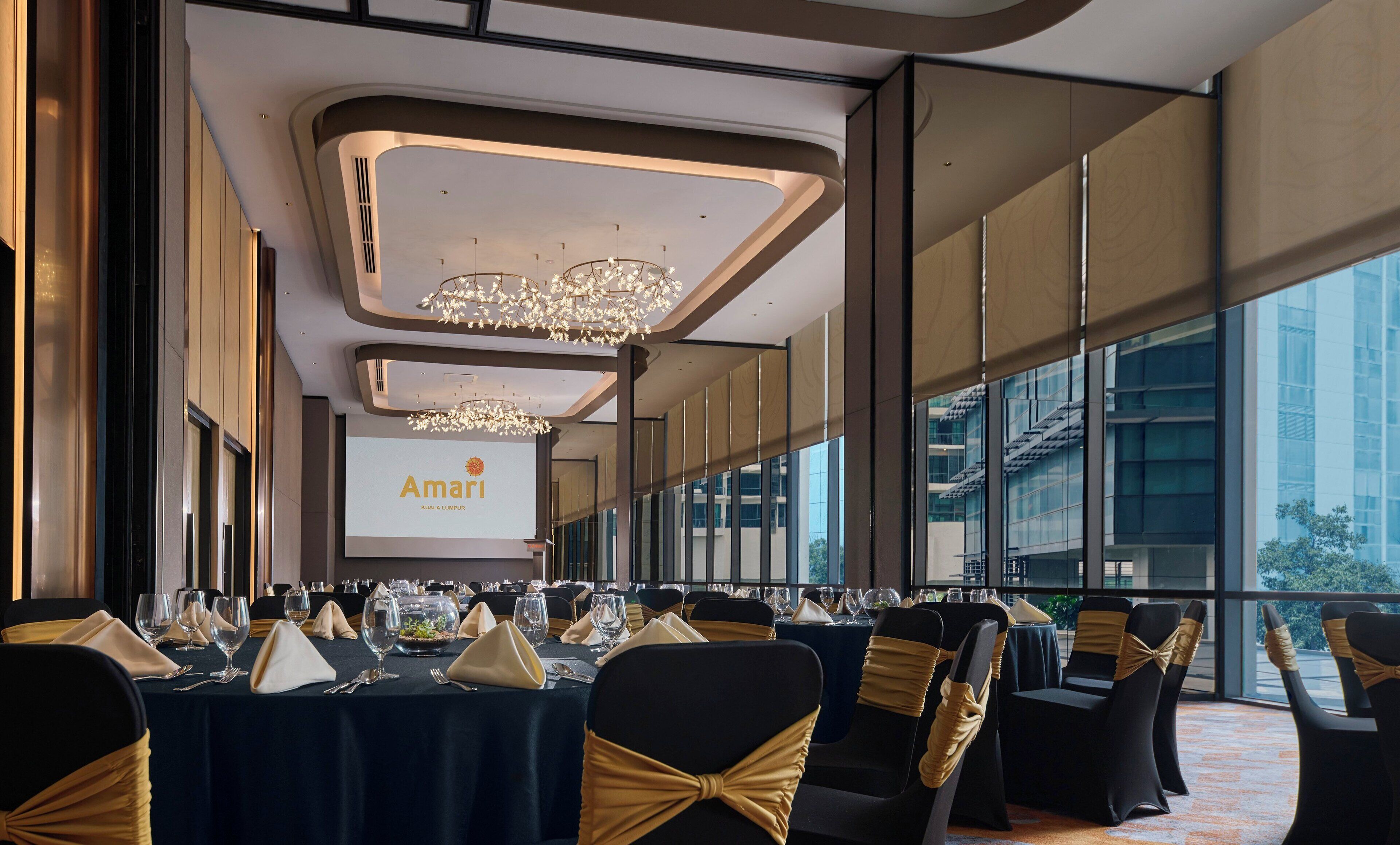 Un salón de conferencias con mesas dispuestas para un evento formal, con una pantalla grande que muestra el nombre 'Amari Kuala Lumpur'.