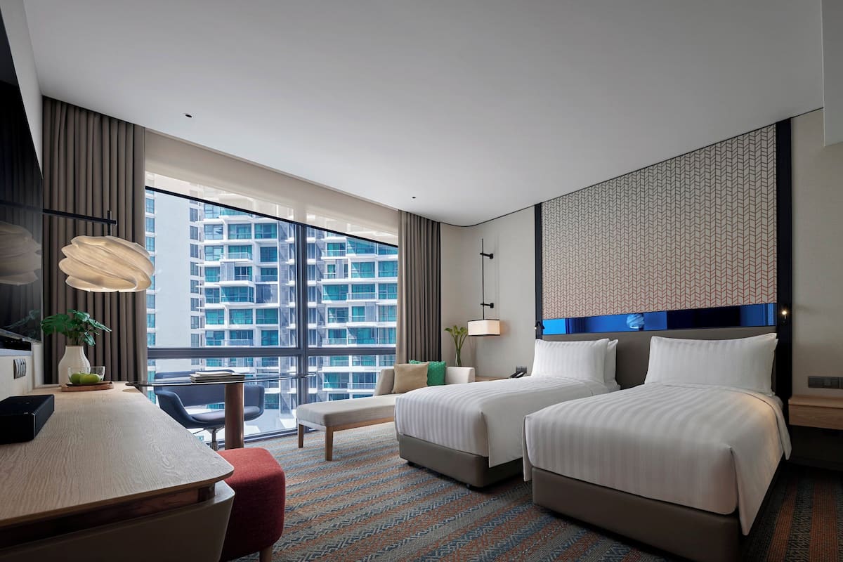A hotel room with two beds, a desk, a chair, a large window with a city view, and a patterned wall.