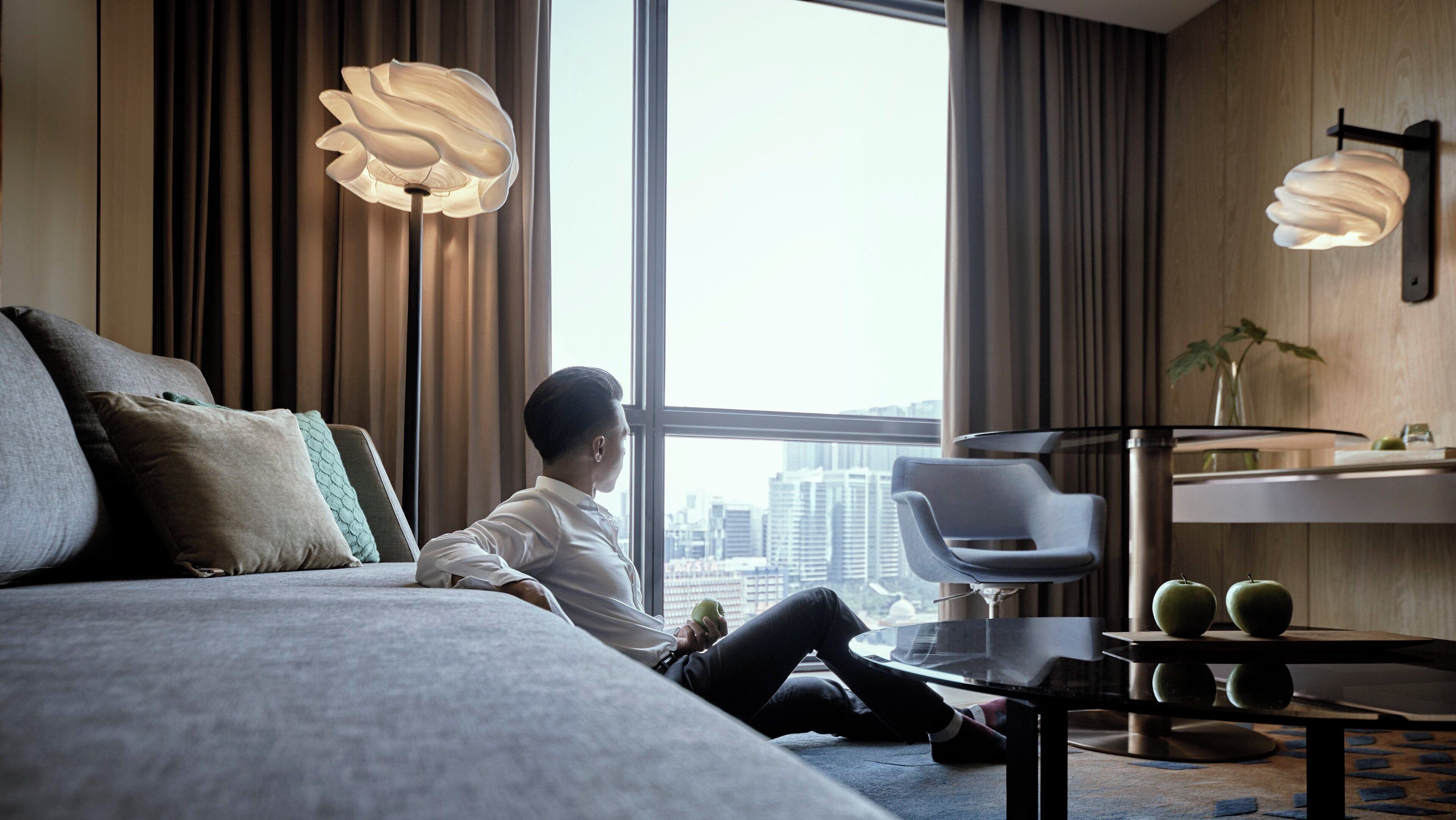 A modern hotel room with a large bed, a sofa, a dining table, and a view of the cityscape through large windows.