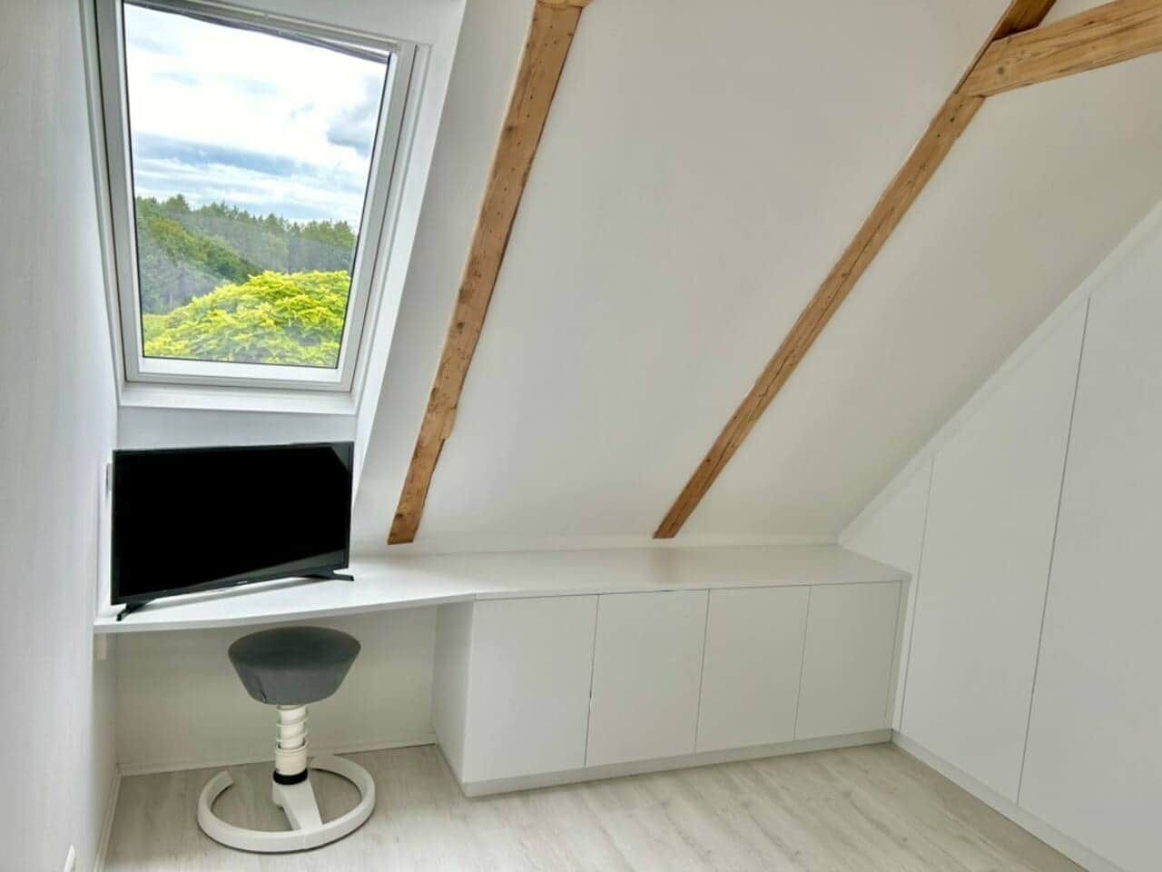 Ein Zimmer mit schrägen Wänden, einem Fenster mit Blick auf Grünflächen, einem weißen Schreibtisch und einem Stuhl.
