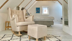 Camera da letto in stile loft con soffitto alto, travi a vista in legno, una grande finestra, un divano, una poltrona e un poggiapiedi.