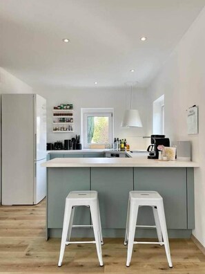A modern kitchen with a central island, two bar stools, a refrigerator, and a window providing natural light. - StylishHomes NEST - A Cosy Adventure (Frauental an der Lassnitz)