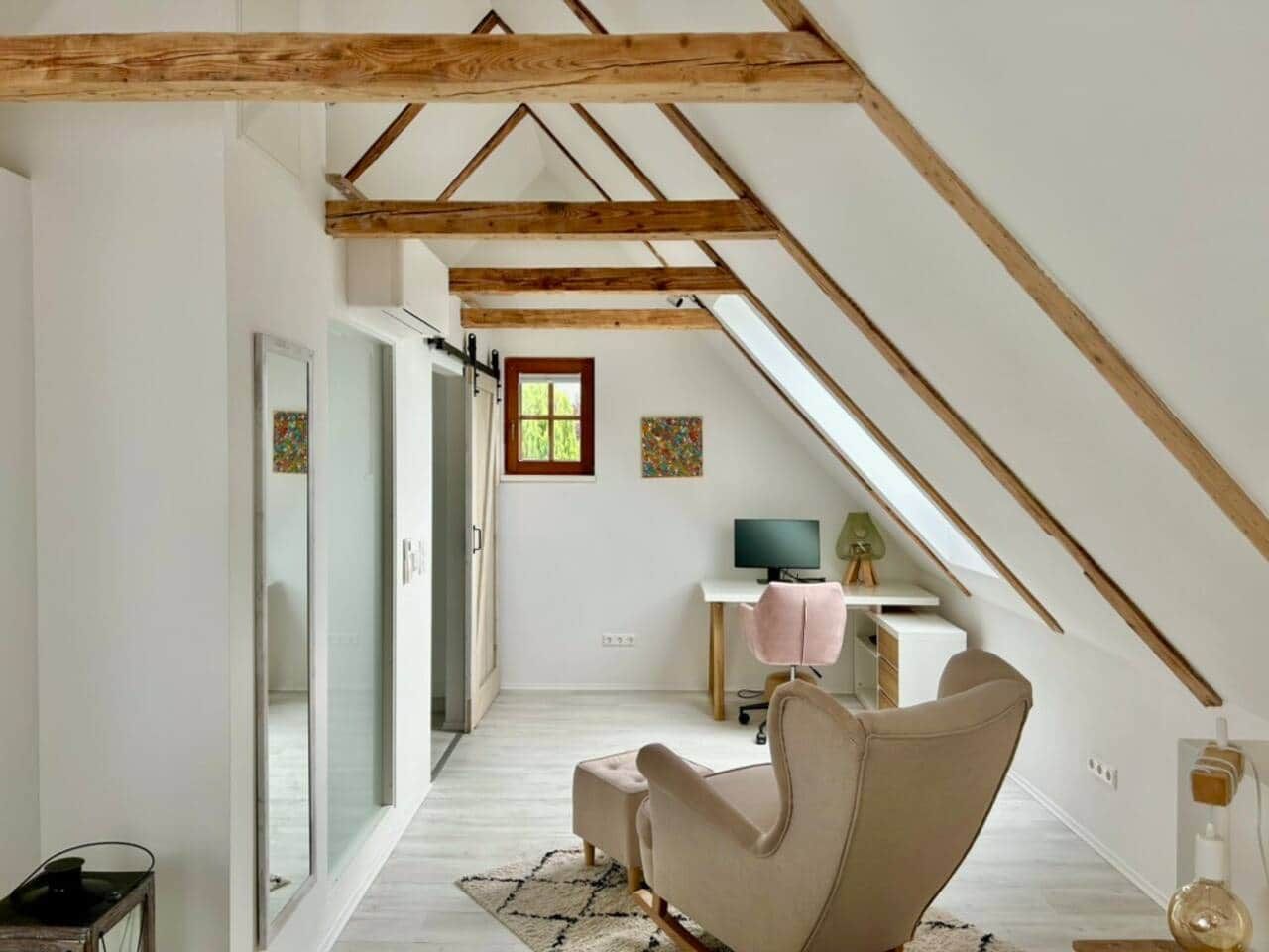 A room with a sloped ceiling, a desk with a computer, a chair, and a window.