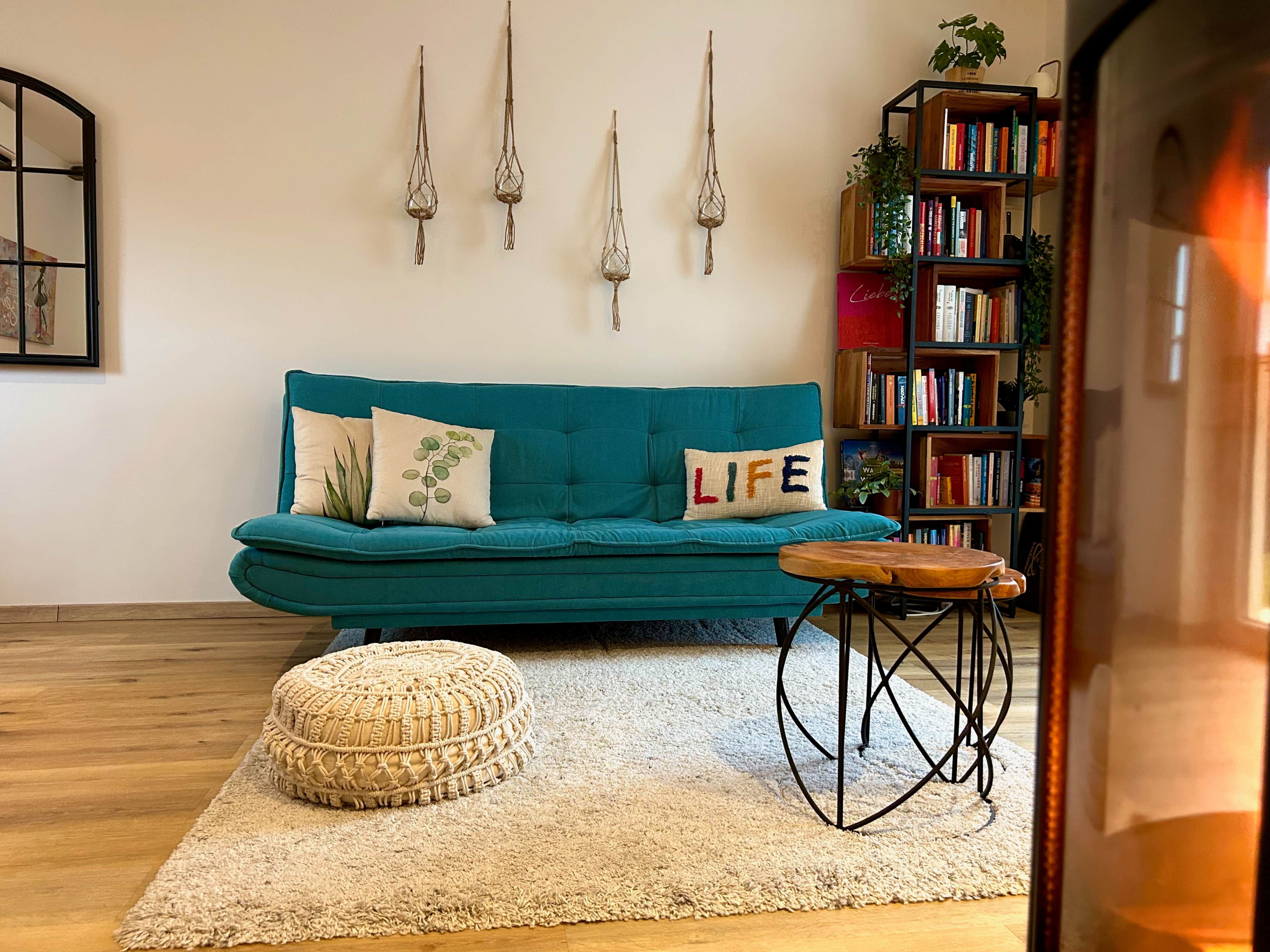 A teal sofa with a 'LIFE' pillow, a round pouf, a small wooden table, a bookshelf, and three hanging light fixtures.