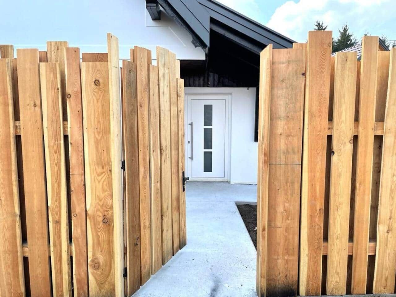 Ein hölzernes Tor führt zu einem Gebäude mit einer weißen Tür und einem Betonweg.