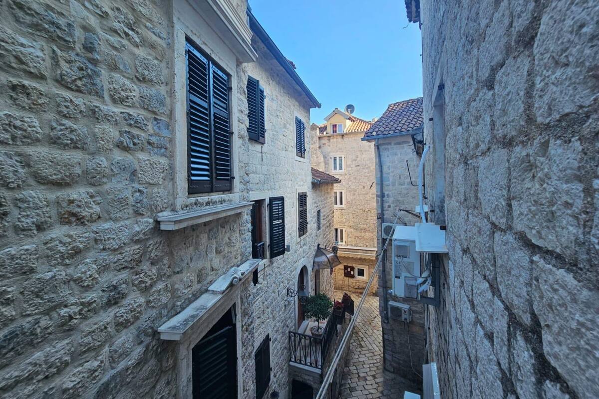 A narrow stone alleyway between buildings with shutters, a small balcony, and a tiled roof visible in the distance.