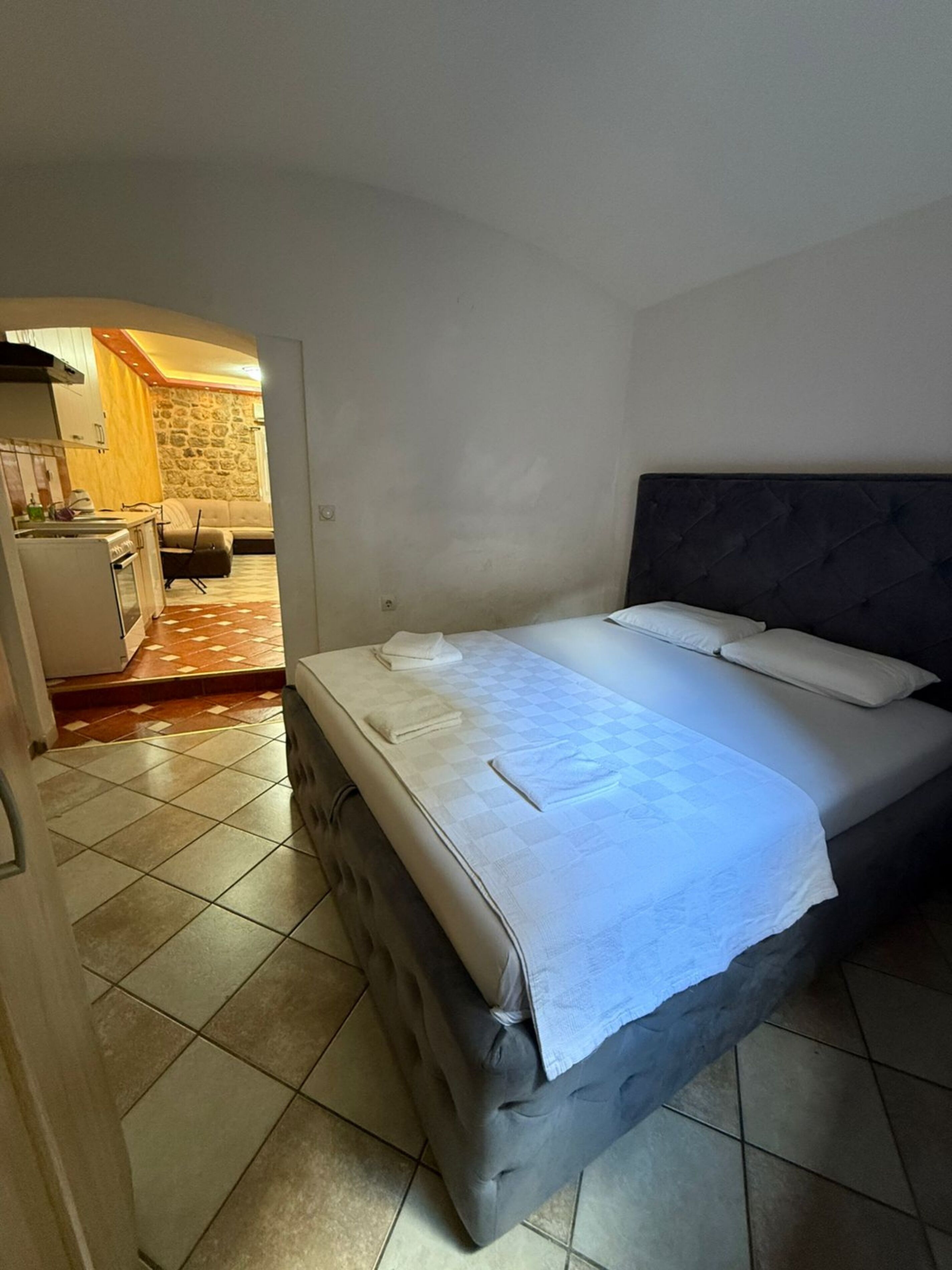 Un dormitorio con cama, cabecera y una zona de cocina visible a través de una puerta abierta.