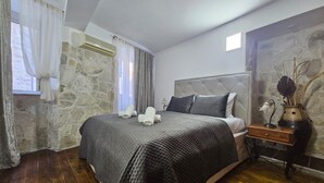 A bedroom with a stone wall, a bed with a grey quilt, a bedside table with a vase, and a window with curtains. - Athos Guesthouse (Kotor)